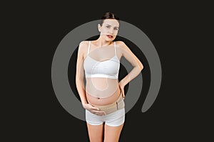 Portrait of pregnant woman in underwear with bandage against backache at black background with copy space. Mother is suffering