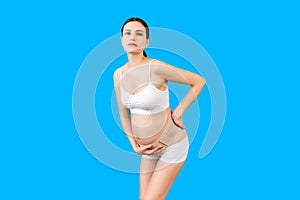 Portrait of pregnant woman in underwear with bandage against backache at blue background with copy space. Mother is suffering from