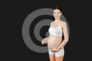 Portrait of pregnant woman in underwear dressing elastic maternity band at black background with copy space. Orthopedic abdominal