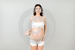 Portrait of pregnant woman in underwear dressing orthopedic bandage to reduce pain in the back at gray background with copy space