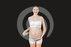 Portrait of pregnant woman in underwear dressing pregnancy corset on the third trimester at black background with copy space.