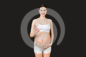 Portrait of pregnant woman in underwear on the third trimester wearing pregnancy belt and showing okay sign at black background.