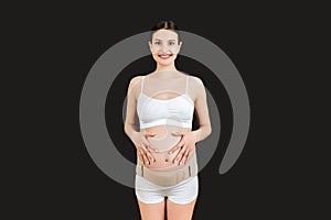 Portrait of pregnant woman in underwear wearing elastic bandage against pain in the back at black background with copy space.