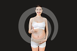 Portrait of pregnant woman in underwear wearing maternity belt on her belly and showing thumb up gesture at black background.