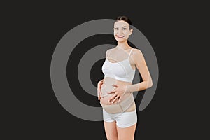 Portrait of pregnant woman in underwear wearing maternity belt to reduce pain in the back at black background with copy space.