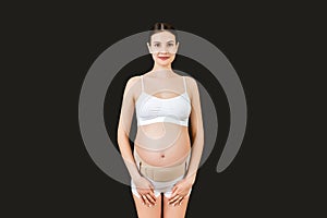 Portrait of pregnant woman in underwear wearing pregnancy bandage at black background with copy space. Orthopedic abdominal