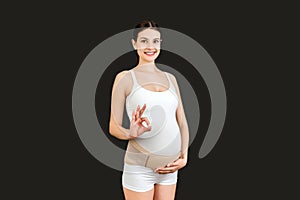 Portrait of pregnant woman wearing pregnancy bandage on her belly to make the backache go away and showing okay gesture at black