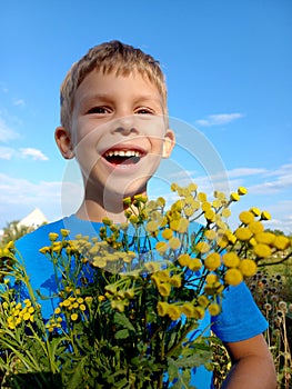 Portrait of preschool boy with bouquet yellow wild flowers against sky. happy child in blue t-shirt holds bouquet of tansy in his