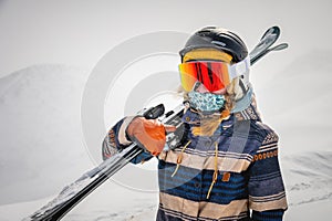 Portrait of a pretty and active woman skier, wearing a mask and holding skis in her hands, active winter holidays