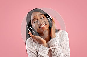 Portrait of pretty African American woman wearing headphones and listening to beautiful music on pink studio background