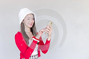 Portrait of a pretty Asian teen girl wearing a red dress and white hat happily using a smartphone on a white background