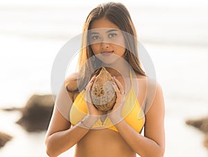Portrait of pretty Asian woman holding coconut and wearing yellow swimsuit. Spending time on the beach. Tanned skin. Long hair.