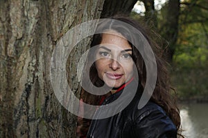 Portrait of a pretty, attractive woman in a leather blouson leaning against a tree trunk