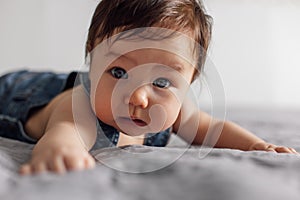 Portrait of pretty baby wearing denim romper lying on gray blanket at home. Scared blue eyed infant child crawl on bed