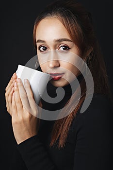 Portrait of pretty beautiful young woman wearing black sweater isolate over dark background. Holding white cup mug. Coffee or tea