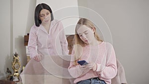 Portrait of pretty blond Caucasian girl using smartphone and listening to adult woman yelling at the background. Teenage