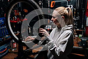 Portrait of pretty blonde female cycling repairman checking bicycle wheel spoke with bike spoke key in repair workshop