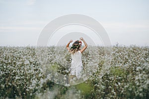 Portrait of a pretty blonde girl with blue eyes with a wreath of flowers on her head walking in field with white flowers