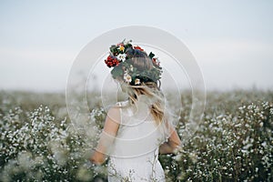 Portrait of a pretty blonde girl with blue eyes with a wreath of flowers on her head walking in field with white flowers