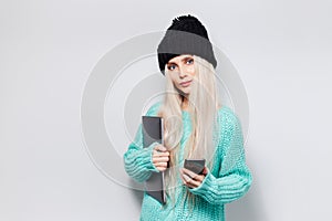 Portrait of pretty blonde girl with laptop in hands using smartphone on white background. Wearing black hat and blue sweater.