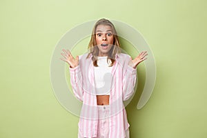 Portrait of pretty caucasian female model with blond short hairstyle, wearing pink outfit, looking amazed, raising hands