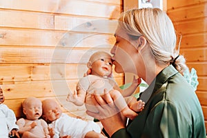 Portrait pretty caucasian young woman kissing realistic newborn baby doll sitting near collection of handmade kids toys