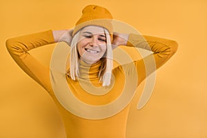 Portrait of pretty and charming young woman with blonde hair wearing yellow clothes, smiling and narrowing her eyes with hands on