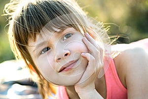 Portrait of pretty child girl with gray eyes and long fair hair leaning on her hands smiling happily outdoors on blurred bright