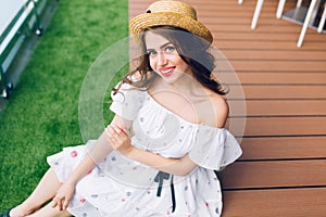 Portrait of pretty girl with long hair in hat sitting on the wood floor outdoor. She wears a white dress with nake