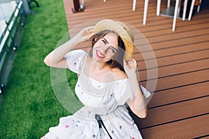Portrait of pretty girl with long hair in hat sitting on the wood floor outdoor. She wears a white dress with nake