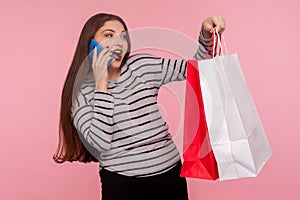 Portrait of pretty happy woman in striped sweatshirt holding shopping bags and enthusiastically talking on mobile phone