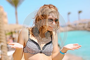 Portrait of pretty happy young woman wearing sunglasses and swimsuit posing near the pool in sunlight. The girl enjoyed her vacati