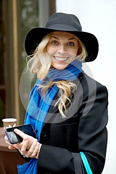 Portrait of pretty lady in lblack hat with cardboard cup and phone in hand