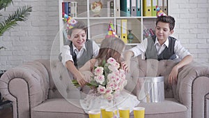 Portrait of pretty little caucasian girl in party hat receiving bouquet of roses and gift box from twin brothers. Happy