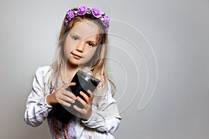 Portrait of pretty little girl with black cup in white dress and purple wreath on gray  background.