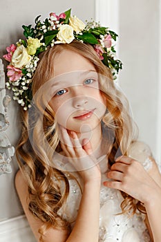 Portrait of a pretty little girl with blond hair in a pink dress is sitting in a bright studio with a wreath of fresh flowers on