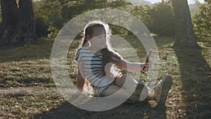 Portrait of pretty little girl in a striped dress laying on green grass in park navigating on a smartphone. Summer