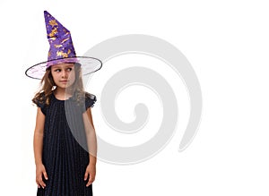 Portrait of pretty little girl wearing a wizard hat and dressed in stylish carnival dress, looking at camera posing with crossed