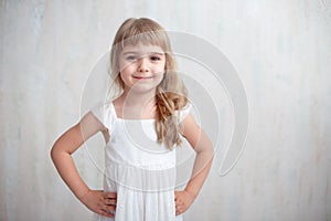 Portrait of pretty little girl in white dress , looking at camera and smiling, standing against gray background