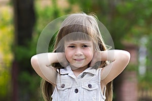 Portrait of a pretty little long-haired blond preschool child girl in sleeveless white dress with hands behind her head smiling