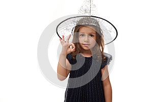 Portrait of pretty little witch girl wearing a wizard hat and dressed in stylish carnival dress, gesturing, showing OK sign with