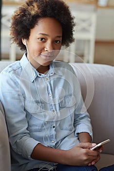 Portrait of pretty mixed race teen girl in earphones looking at camera while sitting on the couch and using mobile phone