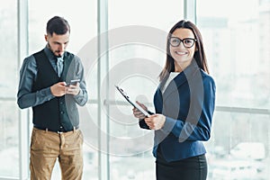 Portrait of pretty office female employee with folder in hands, smiling lovely at the camera and her coleague surfing in