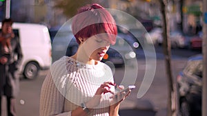 Portrait of pretty pink-haired girl warking with smartphone and turning to camera on blurred street background.