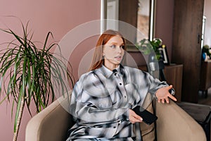 Portrait of pretty red-haired young woman using smartphone sitting on soft armchair at home, looking away. Thoughtful