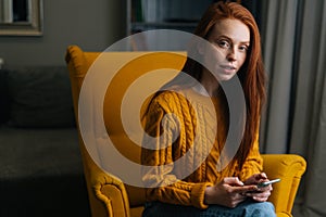 Portrait of pretty red-haired young woman using smartphone sitting in yellow armchair, confident looking at camera.
