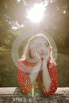 Portrait of pretty redhead woman in red summer loose shirt leaning by hands on a dry beam in wood and posing on a camera