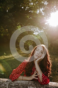 Portrait of pretty redhead woman in red summer loose shirt leaning by hands on a dry beam in wood and posing on a camera