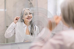 Portrait of pretty senior gray haired woman looking to mirror, cleansing her ears with a cotton swab in modern light