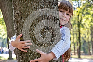 Portrait of a pretty smiling child girl in a paper crown on her head embracing a tree trunk in summer park. Love and care about